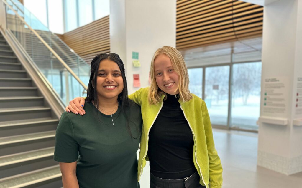 Two women standing in a lobby in front of stairs smiling at the camera.