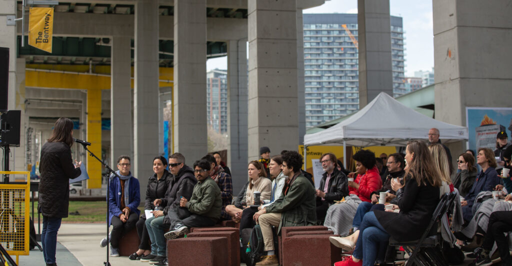 An individual speaking into a microphone in front of a crowd of participants sitting on chairs, underneath the Gardiner Expressway.