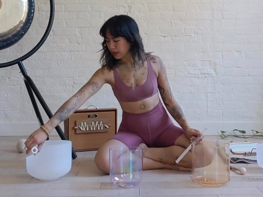 Young woman wearing purple yoga top and shorts sitting cross-legged on the floor. She is holding two tuning instruments with three glass bowls in front of her.