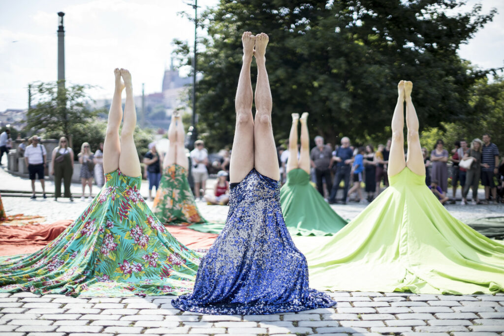 Five people doing headstands on a public street with onlookers gathered in the background.