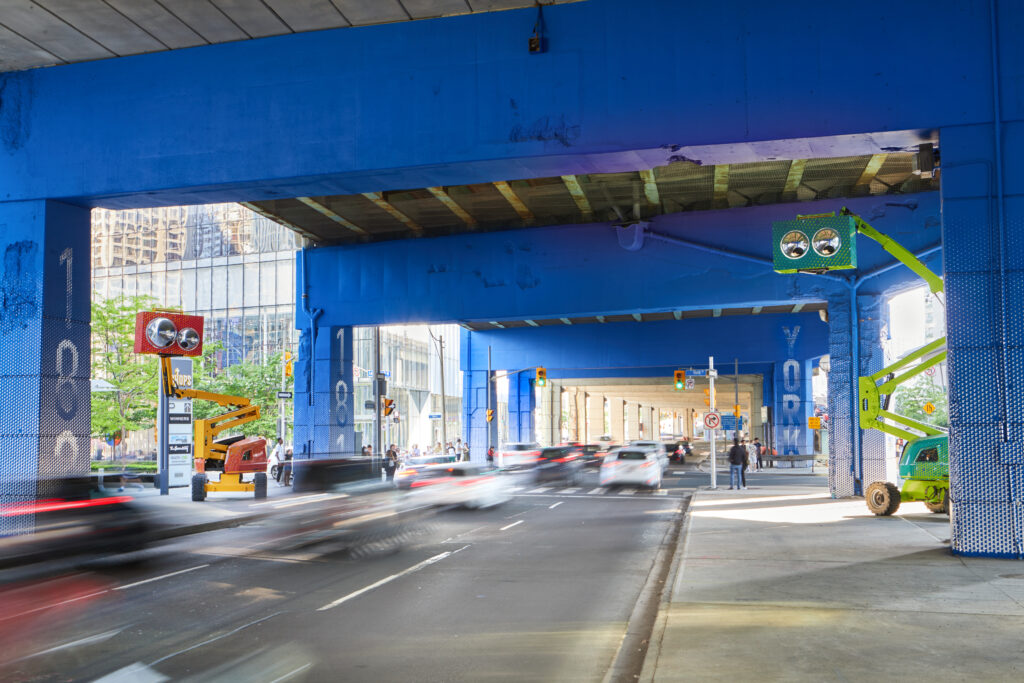 An intersection under an urban highway is painted blue; maintenance equipment have been converted into bright, friendly characters with "googly eyes" overlooking passers-by