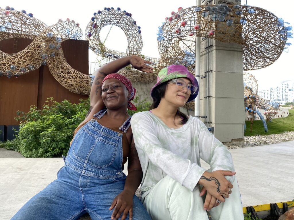 Two people smiling and sitting next to each other on a large rock. Behind them is a building with a large weaving sculpture in front of it.