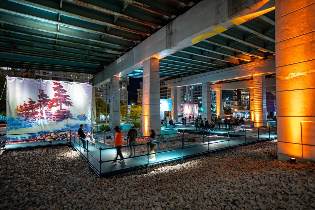 People walking on built pathways underneath a highway at night. Highway columns and large-scale screen installations showing abstract are brightly lit.