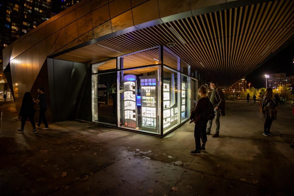 People looking at a shadow puppet display of condos behind a glass window.