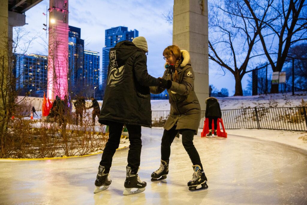 Two people wearing winter gear and skates on ice. One person is guiding another to skate.