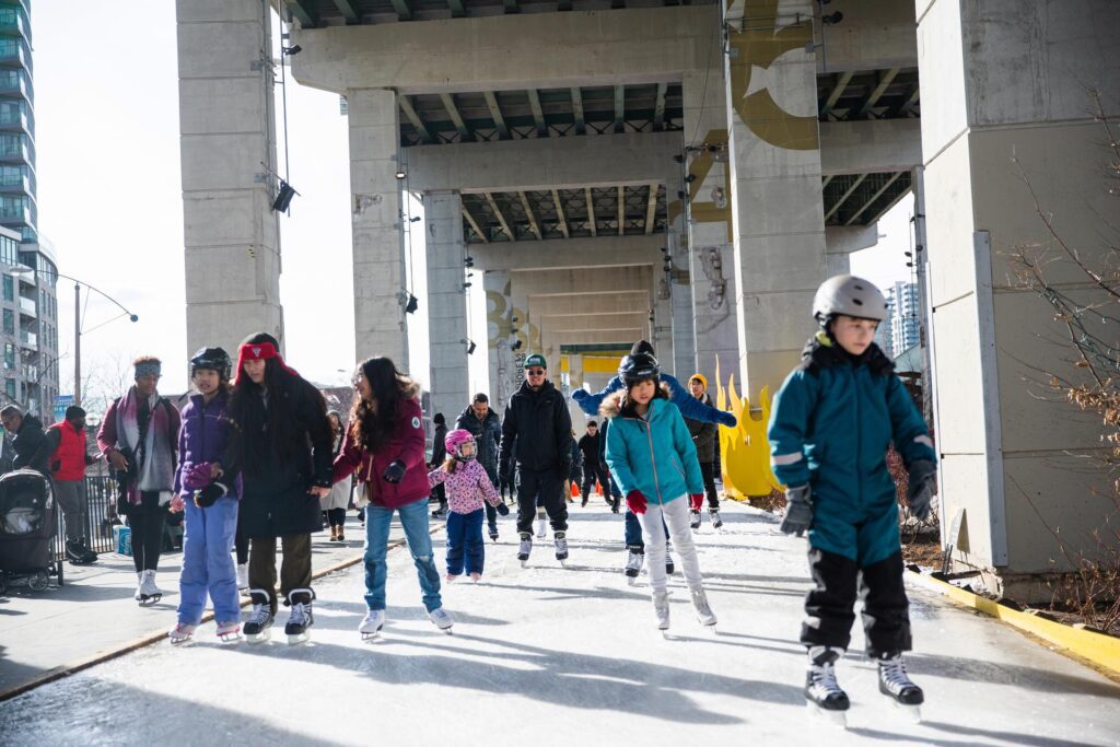 Several younger children and families skating in winter on ice underneath the Gardiner Expressway.