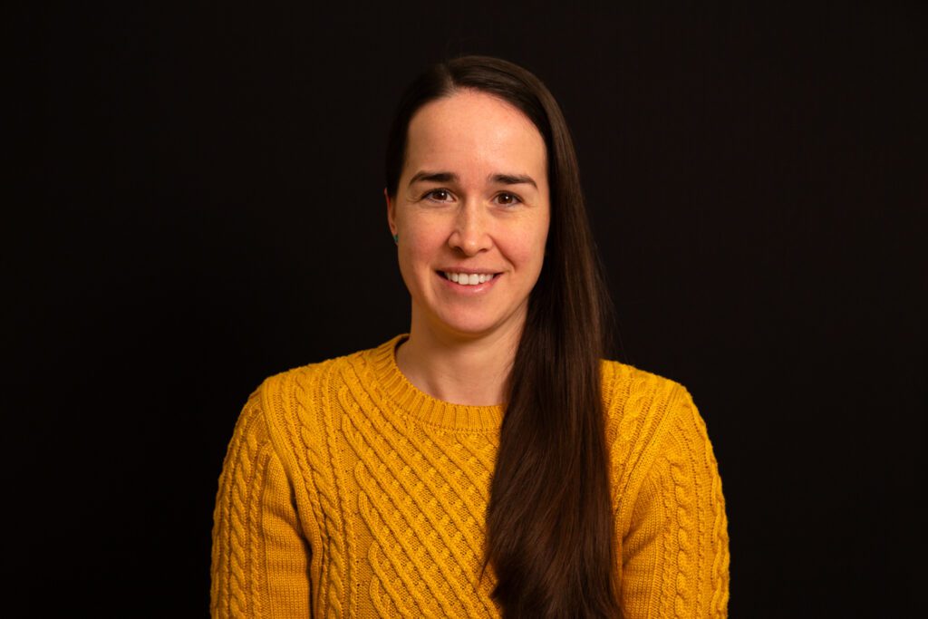 Woman with long brown hair wearing a yellow ochre sweater against a black background.