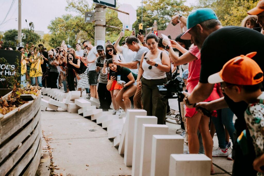 Crowd of people watching and documenting line of giant dominoes falling. 