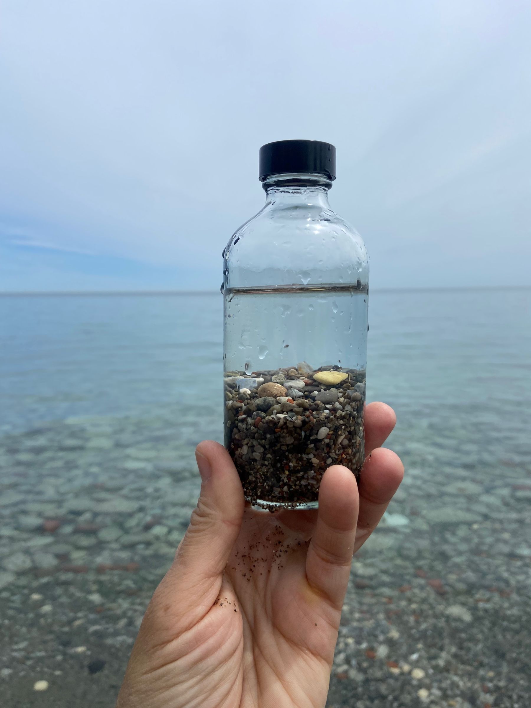 Hand holding a small glass bottle filled with water and pebbles from Lake Ontario, with lake visible in background.