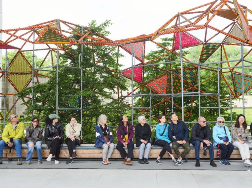 Line of people sitting on a bench in front of a large, sculptural art installation from The Bentway's Softer City exhibition.