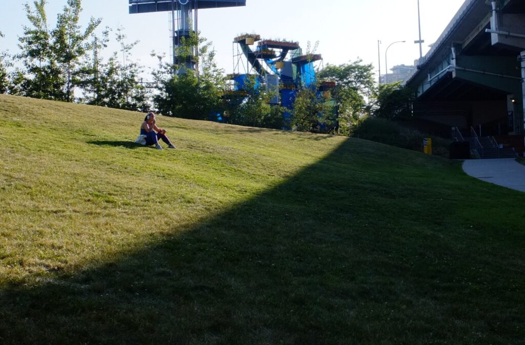 Woman sitting alone on a sunlit grassy hill at The Bentway, near the edge of shade cast by the Gardiner Expressway overhead.