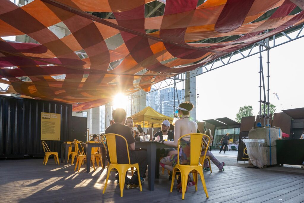 People sitting at tables on a deck, underneath a bright orange and red canopy. 
